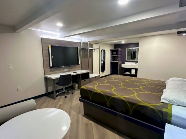 The image shows a modern hotel room with a bed, flat-screen TV, desk, and a table with chairs. A sink and mirror are in the corner.