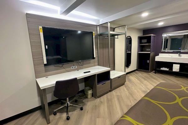 The image shows a modern hotel room with a desk, chair, large TV, open closet, sink, mirror, and a bed.