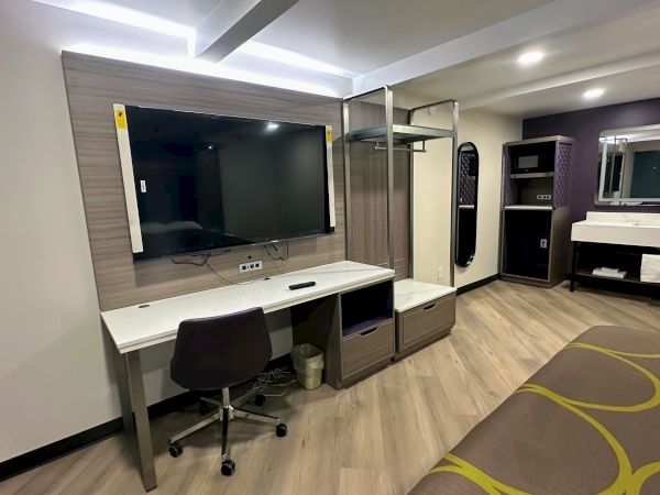 The image shows a modern hotel room with a desk, chair, large TV, open closet, sink, mirror, and a bed.