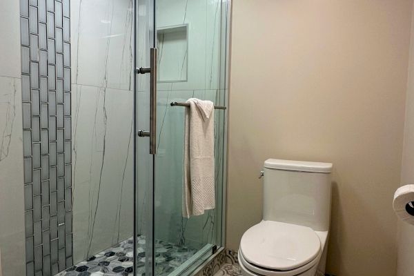 A modern bathroom with patterned floor tiles, a glass shower door, and a toilet. There's a towel on the shower handle and on the floor.