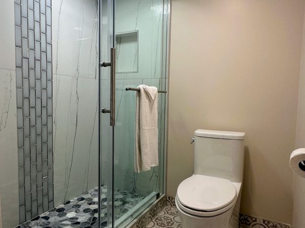 A modern bathroom with patterned floor tiles, a glass shower door, and a toilet. There's a towel on the shower handle and on the floor.