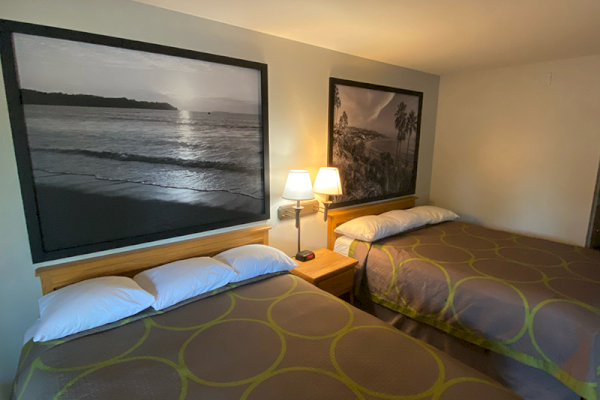 A hotel room with two double beds, each featuring two pillows, a nightstand with lamps between them, and framed landscape photos on the walls.