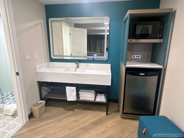 A hotel room corner with a sink, mirror, microwave, mini-fridge, and bathroom entryway.