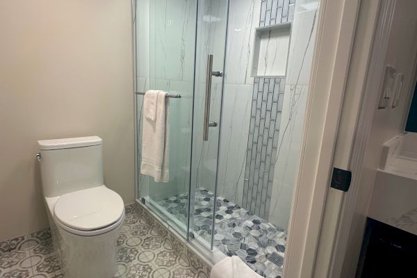A modern bathroom with a patterned floor, toilet, glass shower enclosure, and a white towel hanging on the door.