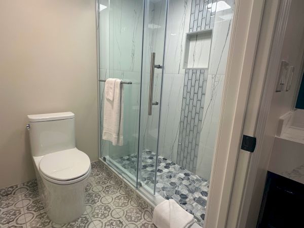 A modern bathroom with a patterned floor, toilet, glass shower enclosure, and a white towel hanging on the door.