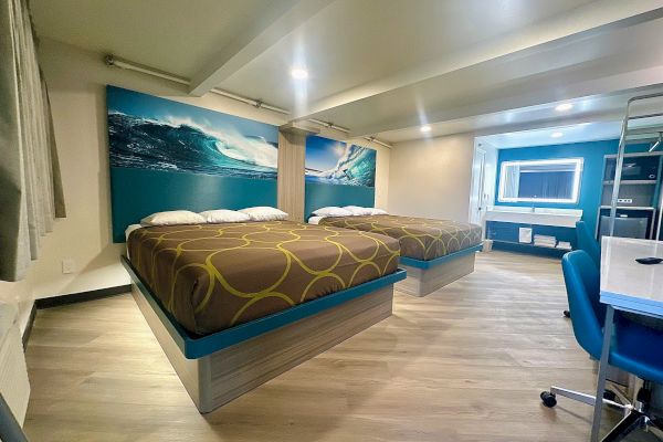 A hotel room features two beds with ocean-themed artwork, a desk chair, and a neatly arranged vanity area under bright lighting.