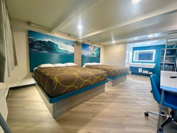 A hotel room features two beds with ocean-themed artwork, a desk chair, and a neatly arranged vanity area under bright lighting.