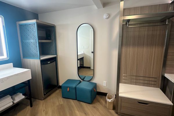 A hotel room corner with a mirror, two teal ottomans, a small fridge, microwave, sink, towels, and wooden floors.
