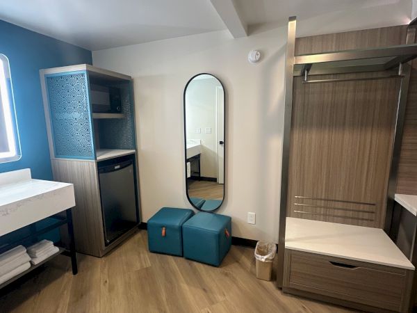 A hotel room corner with a mirror, two teal ottomans, a small fridge, microwave, sink, towels, and wooden floors.