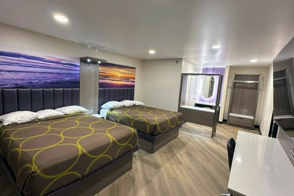 The image shows a modern hotel room with two beds, a large beach art piece, desk, mirror, and wooden floor, creating a cozy atmosphere.