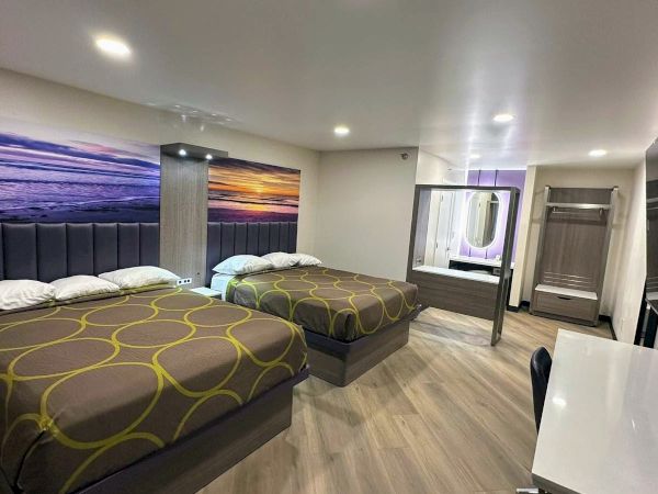 The image shows a modern hotel room with two beds, a large beach art piece, desk, mirror, and wooden floor, creating a cozy atmosphere.