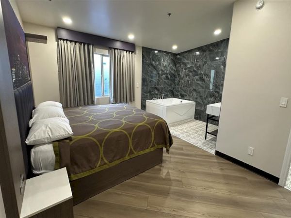 A modern hotel room with a king bed, white bedding, circular-pattern bedspread, a freestanding bathtub near a dark marble wall, and a small desk area with a window and curtains.
