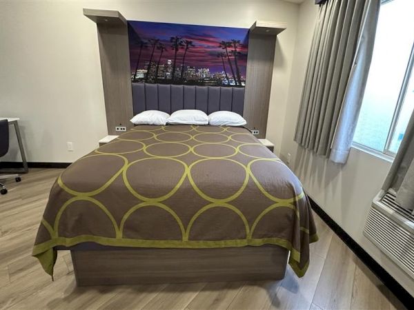 A tidy hotel room with a bed featuring a circular-patterned brown and olive bedspread, a headboard with a sunset mural, and a desk by the window.
