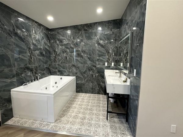 A modern bathroom with a white corner bathtub, dark marble walls, patterned floor tiles, and a white sink with a mirror, all neatly arranged.